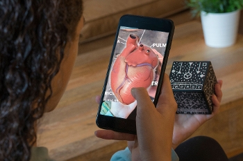 over the shoulder view of girl holding MERGE augmented reality cube in left hand, viewing it through a smartphone held in right hand. Smartphone screen shows image of a human heart