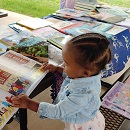 A child reading a book on an outdoor picnic table covered with children's books. Part of a "Back to Nature" outdoor program.